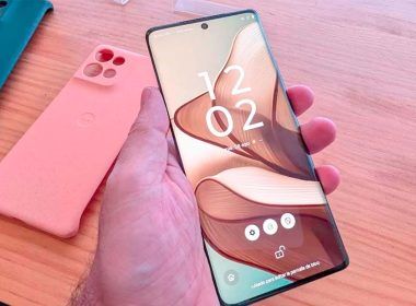 Primer plano de la pantalla de un smartphone Motorola encendido junto a dos fundas protectoras sobre una mesa de madera.