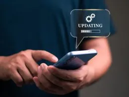 Primer plano de unas manos sosteniendo un smartphone mientras se muestra una burbuja gráfica con la palabra UPDATING, un icono de engranajes y una barra de carga.
