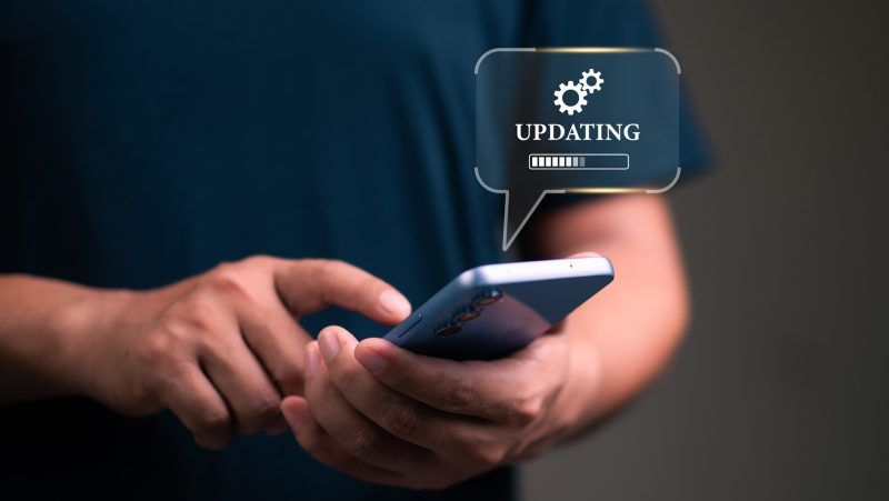 Primer plano de unas manos sosteniendo un smartphone mientras se muestra una burbuja gráfica con la palabra UPDATING, un icono de engranajes y una barra de carga.