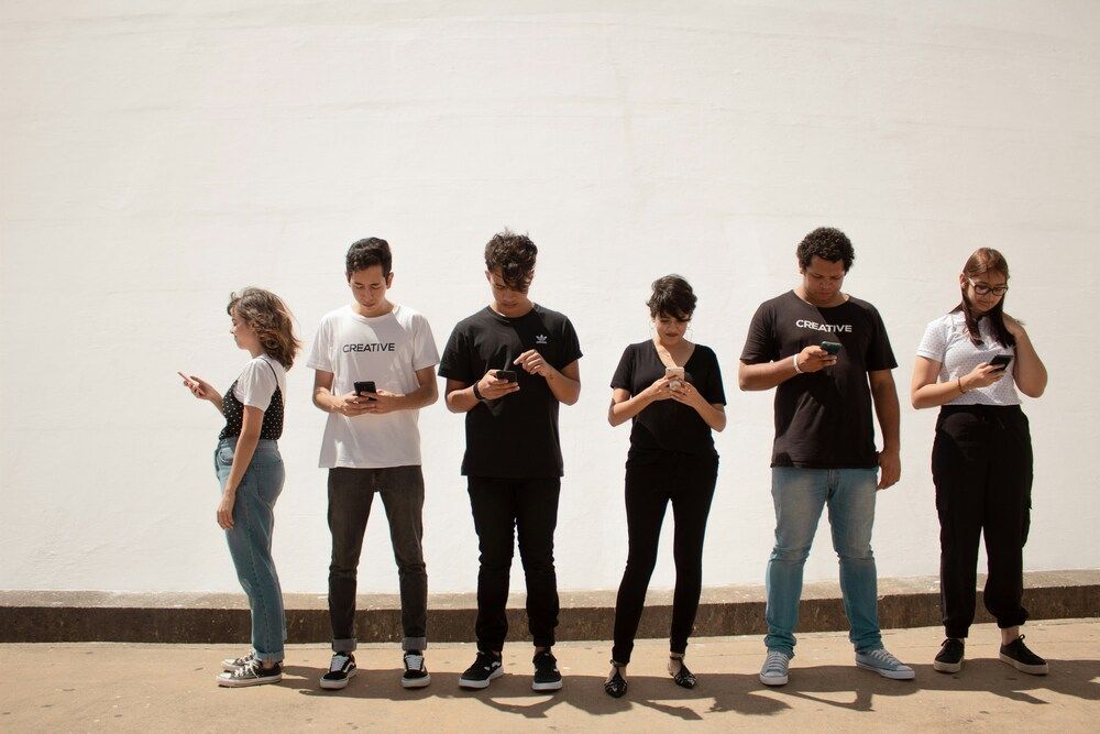 Seis personas jóvenes en fila mirando sus teléfonos móviles frente a una pared blanca.