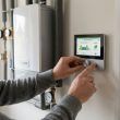 Persona ajustando la configuración de temperatura en un termostato digital de pared junto a una caldera doméstica.