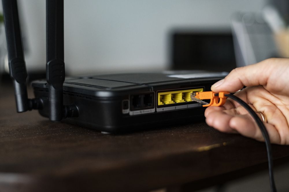 Primer plano de una mano conectando un cable Ethernet naranja en el puerto LAN amarillo de un router inalámbrico negro.