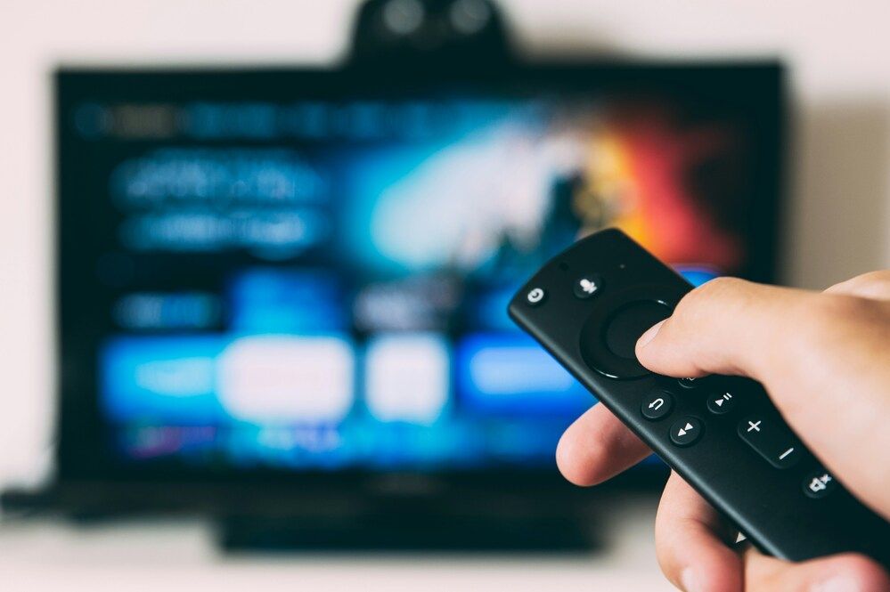 Primer plano de una mano sujetando un mando a distancia frente a una Smart TV con la interfaz de usuario desenfocada.