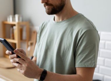 Hombre con barba manejando un teléfono móvil en una cocina con azulejos blancos.