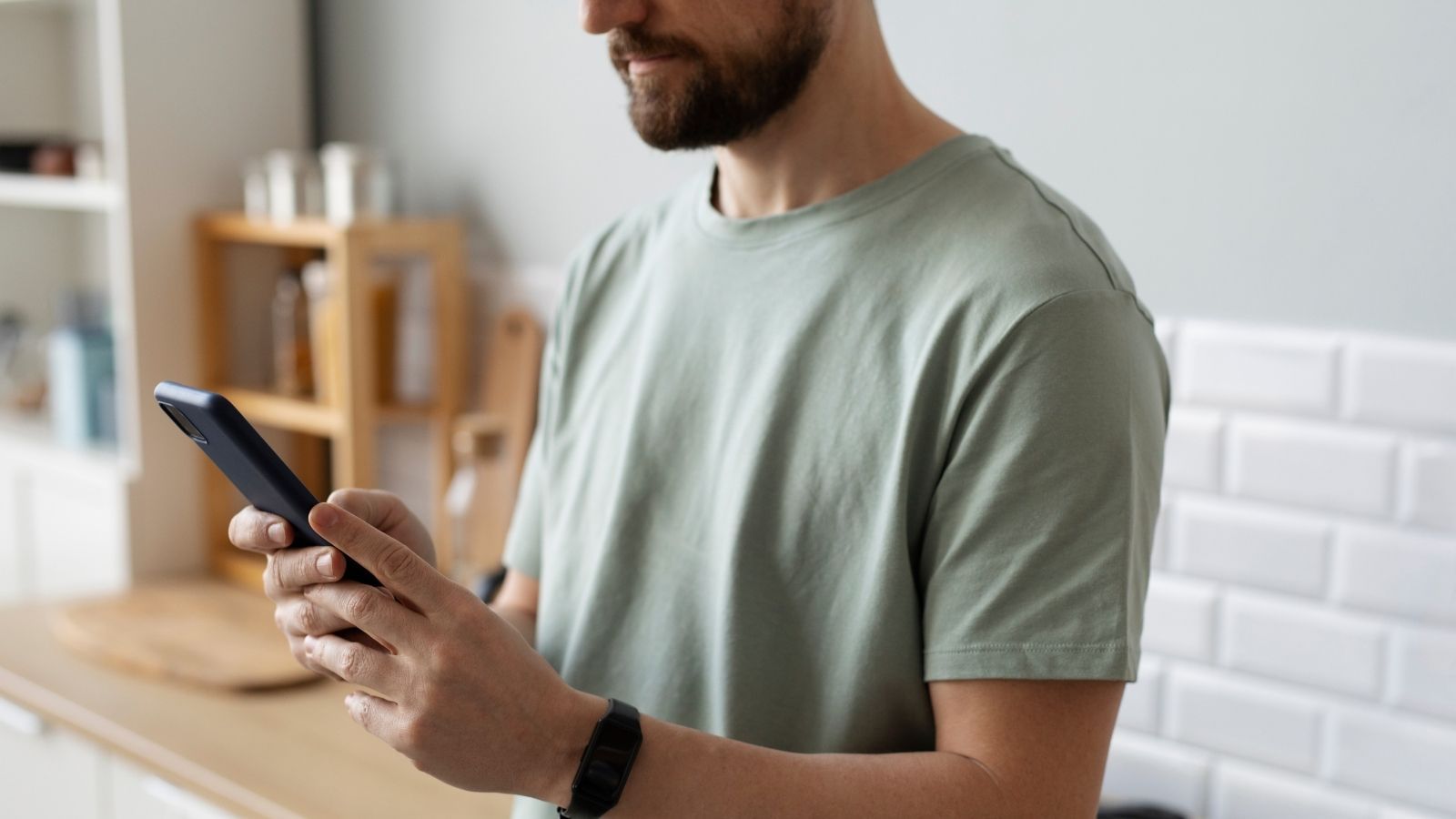 Hombre con barba manejando un teléfono móvil en una cocina con azulejos blancos.