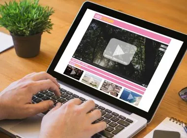 Manos de una persona escribiendo en el teclado de un ordenador portátil que muestra una plataforma de vídeo en streaming sobre una mesa de madera.