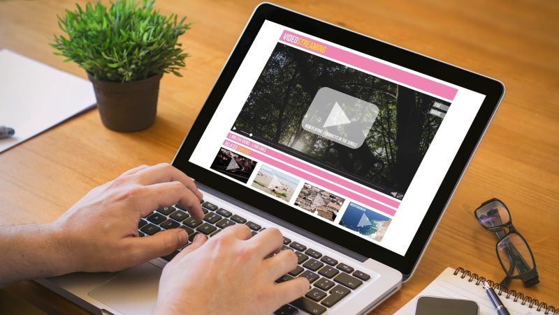 Manos de una persona escribiendo en el teclado de un ordenador portátil que muestra una plataforma de vídeo en streaming sobre una mesa de madera.